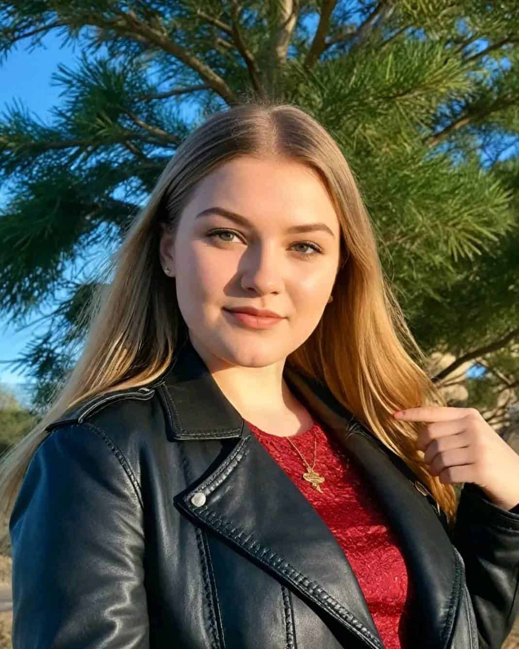 Girl with long blonde hair wearing a black leather jacket, red top, and gold necklace, standing outdoors with a pine tree and blue sky in the background, enjoying a sunny day.