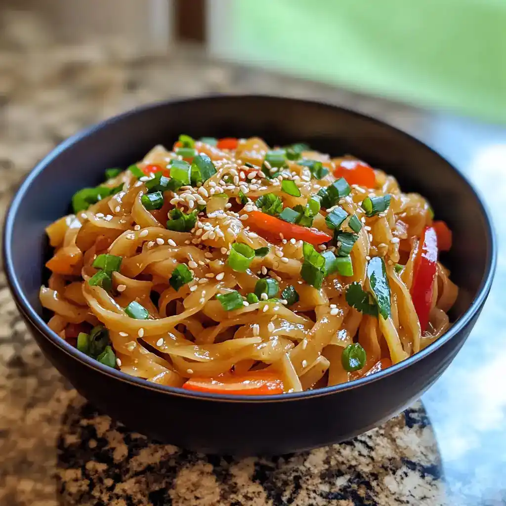 Stir-fried veggie noodles with colorful vegetables, sesame seeds, and green onions served in a black bowl on a kitchen countertop.