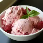 Sweet creamy raspberry ice cream with fresh raspberries and basil garnish. Perfect for summer dessert or homemade treat. Deliciously fruity and refreshing.