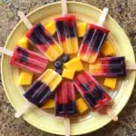 Colorful layered fruit popsicles with mango, blueberry, and berry flavors on a yellow plate. Perfect for summer desserts and healthy treats.