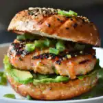 Savory salmon burger with avocado and green onions, grilled bun, black and white sesame seeds, served on a white plate.