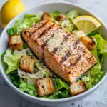 Pan-seared salmon salad with grilled lemon, croutons, and shaved Parmesan cheese, featuring fresh lettuce and a creamy dressing for a healthy, delicious meal.