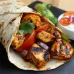 Grilled chicken wrap with fresh vegetables and cilantro, served with a side of yogurt sauce on black slate plate. Perfect for quick, healthy lunch or dinner options.