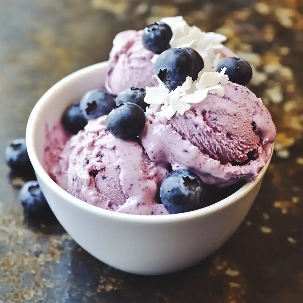 Creamy blueberry ice cream topped with fresh blueberries and white chocolate shavings in a white bowl, perfect for summer desserts and homemade ice cream recipes.