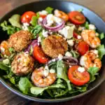 Crab cake salad with fresh vegetables, cherry tomatoes, red onions, shrimp, and feta cheese on a black plate. Healthy, flavorful, and easy-to-make seafood salad recipe from Station Recipes.