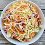 Fresh coleslaw salad with shredded cabbage, carrots, and purple cabbage on a white bowl over rustic wooden table. Perfect for summer BBQs and healthy side dishes.