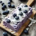 Fresh blueberry and coconut ice cream bar on a wooden tray with scattered blueberries and coconut flakes for a refreshing summer treat.