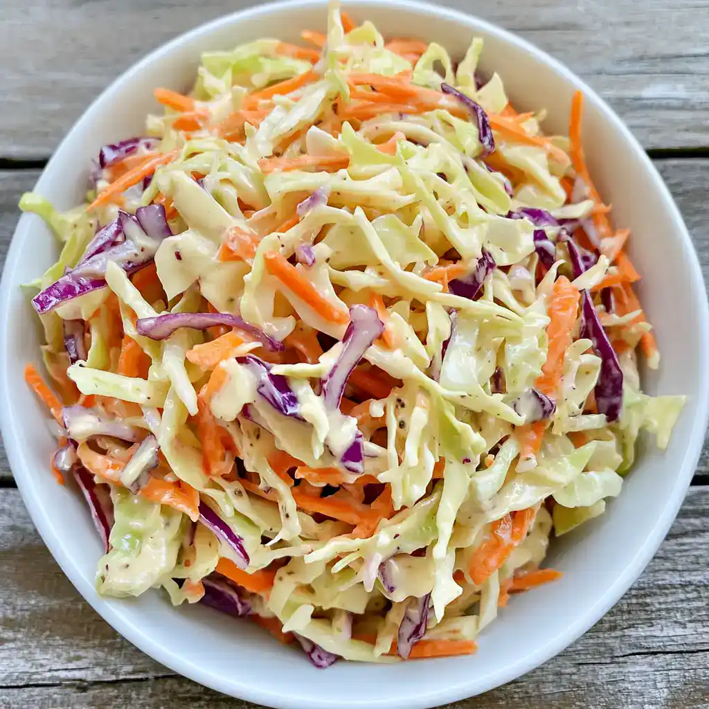 Fresh coleslaw salad with shredded cabbage, carrots, and purple cabbage on a white bowl over rustic wooden table. Perfect for summer BBQs and healthy side dishes.