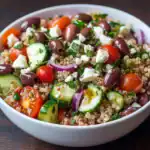 Fresh Mediterranean quinoa salad with cherry tomatoes, cucumbers, Kalamata olives, red onions, crumbled feta cheese, and chopped parsley in a white bowl.