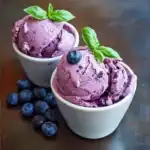 Creamy blueberry ice cream topped with fresh blueberries and basil leaves in white bowls, showcasing a delicious summer dessert. Perfect for blueberry lovers and homemade ice cream recipes.