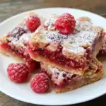 Rich raspberry and almond cake squares with powdered sugar and fresh raspberries on a white plate. Perfect for dessert or special occasions, featuring fruity, nutty flavors.