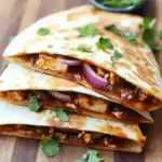 Savory chicken quesadillas filled with melted cheese, beans, and spicy sauce, garnished with fresh cilantro and red onions on a rustic wooden table. Perfect for quick, tasty meals or snacks.