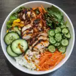 Grilled chicken salad bowl with fresh vegetables, cucumber, carrots, and mixed greens, topped with sesame seeds and savory sauce, perfect for healthy meal options from Station Recipes.