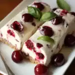 Creamy cherry cheesecake bars topped with fresh cherries and basil leaves on a white serving dish, perfect for summer desserts and cherry recipes.