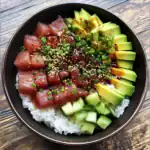 Fresh tuna poke bowl with diced avocado, cucumber, and green onions on a bed of sushi rice, drizzled with soy sauce and sesame seeds, served in a black bowl.