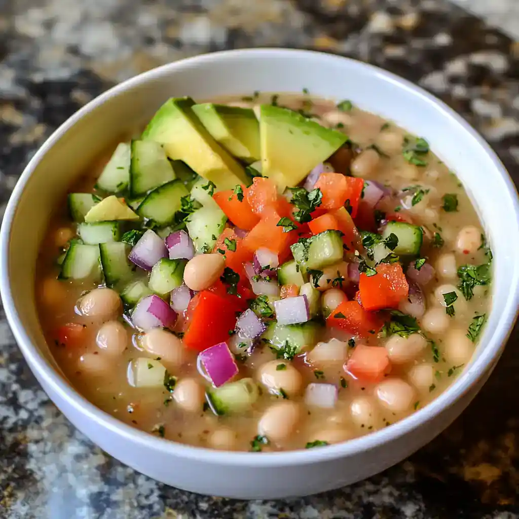 Creamy chickpea salad with fresh vegetables, including cucumber, tomato, red onion, and avocado, garnished with chopped herbs, served in a white bowl on a rustic countertop.