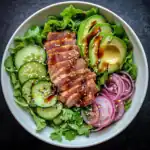 Sliced seared tuna salad with avocado, cucumber, red onion, and sesame seeds on a bed of greens, drizzled with balsamic glaze. Perfect for healthy, quick lunch recipes and fresh seafood dishes.