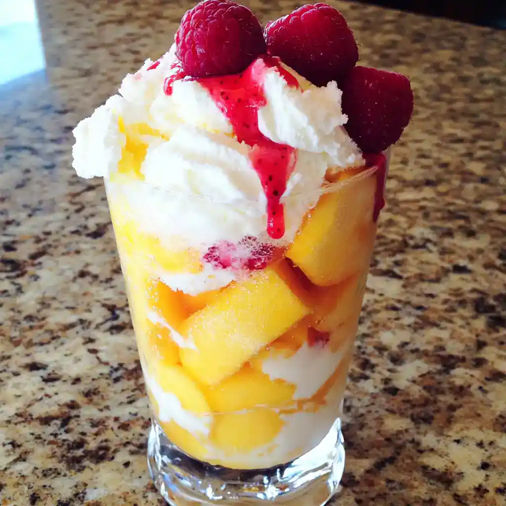 Fresh peach and raspberry parfait with whipped cream, drizzled with berry syrup, served in a tall glass on a granite countertop. Perfect for summer desserts and fruit parfait recipes.