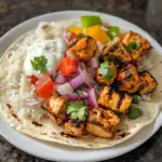 Grilled chicken tikka with rice, fresh vegetables, and creamy yogurt sauce on a soft tortilla, served on a white plate. Perfect flavorful Indian-inspired dish for lunch or dinner.