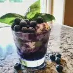 Fresh blueberry dessert parfait with basil leaves, creamy layers, and a dusting of powdered sugar, served in a clear glass on a marble countertop. Perfect for summer or breakfast.