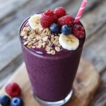 Vegan berry smoothie with fresh raspberries, blueberries, banana slices, and granola topping in a clear glass on rustic wooden background. Perfect healthy breakfast or snack option.