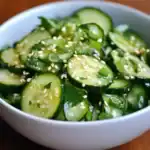 Cucumber salad with sesame seeds, chopped green onions, and fresh parsley in a white bowl, made with sliced cucumbers and light seasoning, ideal for healthy side dishes and summer recipes.