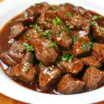 Beef stew with tender chunks of beef in rich gravy garnished with fresh chopped parsley on a white plate. Perfect comfort food recipe for cozy dinner, featuring savory flavors and hearty texture.