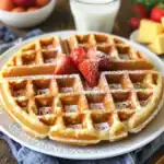 Fluffy Belgian waffles topped with strawberries and powdered sugar on a white plate, perfect for breakfast or brunch, served with fresh fruit, syrup, and whipped cream.