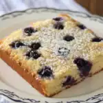 Blueberry sheet cake with powdered sugar on a decorative plate, featuring fresh blueberries and moist crumb. Perfect for desserts or snack recipes.