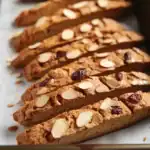 Buttercrunch almond cranberry biscotti on baking sheet with almonds and cranberries for homemade cookie recipe.