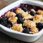 Blackberry cobbler with crumbly topping in a white baking dish, fresh blackberries and berry juice visible, perfect for dessert or summertime treat.