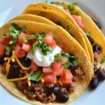 1. Delicious homemade crunchy taco shells filled with seasoned ground beef, black beans, shredded cheese, sour cream, fresh tomatoes, and cilantro on a white plate.