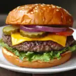 Juicy cheeseburger with pickles, onions, lettuce, tomato, and sesame seed bun on a white plate. Perfect for a classic American meal or summer cookout.