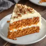Rich carrot cake slice with cream cheese frosting and chopped pecans on top, served on a white plate, perfect for dessert or celebration.