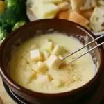 Creamy cheese fondue with melted cheese cubes in a rustic brown bowl, served with fresh vegetables and bread for dipping, perfect for cozy gatherings and festive occasions.