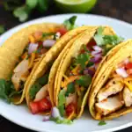 Crispy chicken tacos with fresh vegetables, tomatoes, cilantro, and shredded cheese on a white plate - Station Recipes.