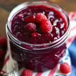 Sweet homemade raspberry jam in a glass jar with fresh raspberries on top, perfect for breakfast or toast.