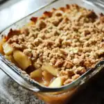 Golden baked apple crumble dessert served in a glass baking dish with a crispy streusel topping. Perfect for fall and winter gatherings, featuring tender apples and a crunchy topping for a cozy treat.