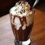 Creamy whipped topping with chocolate shavings on an iced coffee milkshake in a tall glass with chocolate syrup drizzle, served with a metal straw.