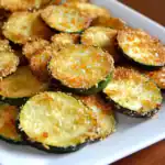 Crunchy baked zucchini chips with golden breadcrumb topping on a white serving platter. Perfect healthy snack or appetizer with crispy texture and cheesy flavor.