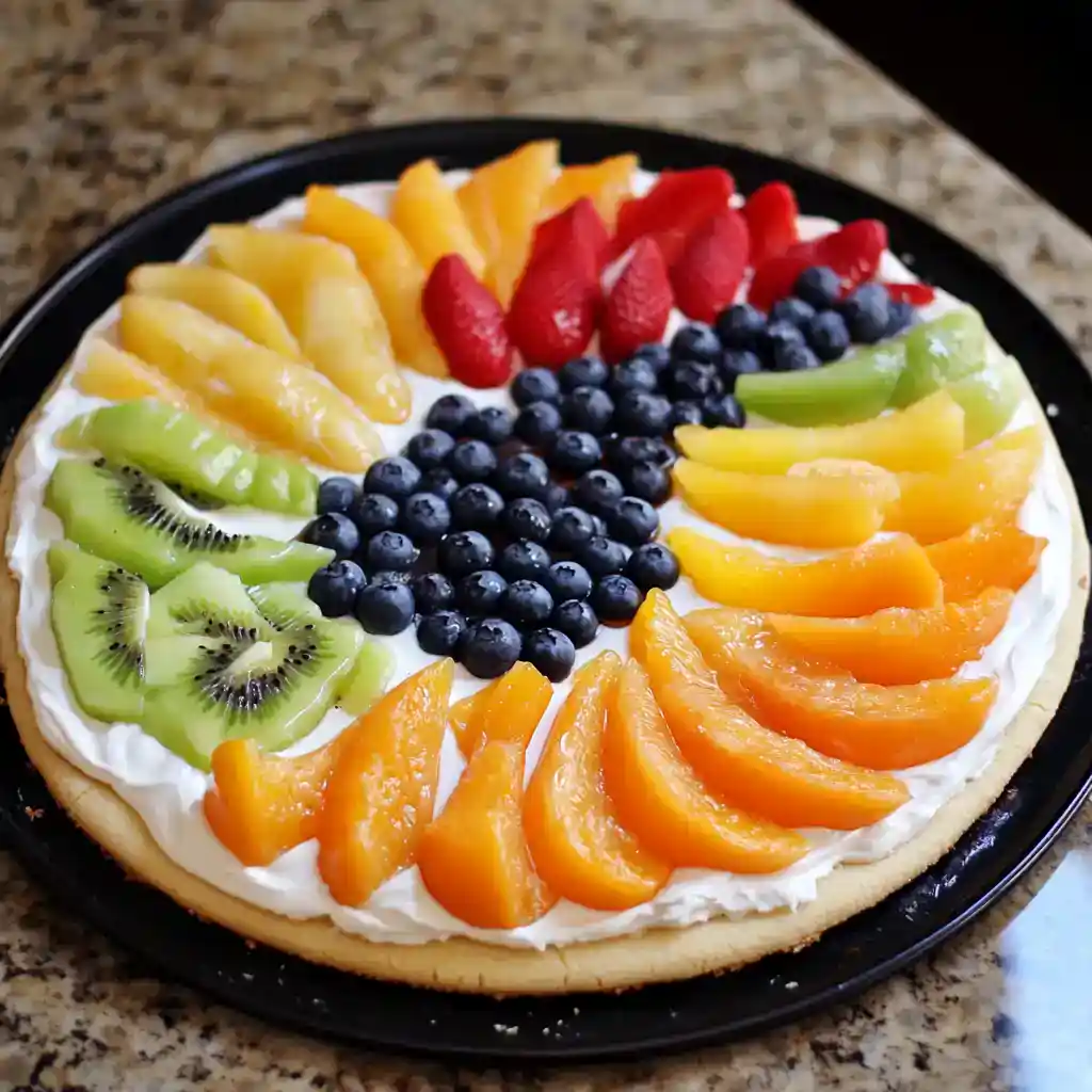 Fresh fruit pizza topped with colorful sliced fruits including kiwi, strawberries, blueberries, peaches, pineapple, and grapes on a crispy cookie crust, layered with creamy whipped topping.