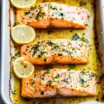 Baked lemon garlic salmon fillets with fresh herbs in a baking dish, garnished with lemon slices, perfect for healthy dinner recipes and seafood lovers.