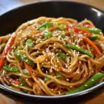 Stir-fried vegetable chow mein with noodles, colorful bell peppers, and green onions garnished with sesame seeds in a black bowl.