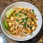 Stir-fried tofu with rice noodles, bean sprouts, chopped peanuts, fresh cilantro, and lime wedges, served in a white bowl. Perfect for a flavorful, vegetarian Asian-inspired meal at home or on the go.