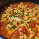 Creamy pasta e fagioli soup with diced tomatoes, carrots, and herbs, topped with fresh parsley and cheese, served hot in a black bowl, highlighting a comforting and hearty homemade recipe.