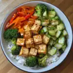 Steamed tofu with sesame seeds, fresh broccoli, cucumber, and sliced carrots served over white rice in a bowl. Healthy vegetarian Asian-inspired meal perfect for quick, nutritious dinners or lunch.