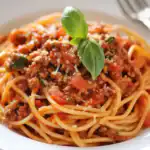 Ground beef spaghetti with tomato sauce and fresh basil on top, served on a white plate, showcasing a classic comfort food dish.