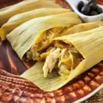 Tamale dish wrapped in corn husk with a side of black olives on a patterned plate.
