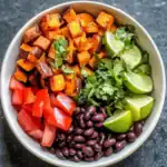 Flavorful sweet potato and black bean bowl garnished with fresh lime, cilantro, and chopped tomatoes for a nutritious meal. Perfect for vegan, gluten-free, and healthy eating enthusiasts.