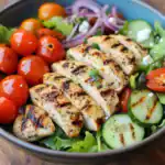 Grilled chicken breast with char marks over a bed of fresh greens, cherry tomatoes, cucumber, and red onions. Ideal for healthy eating and summer meals.